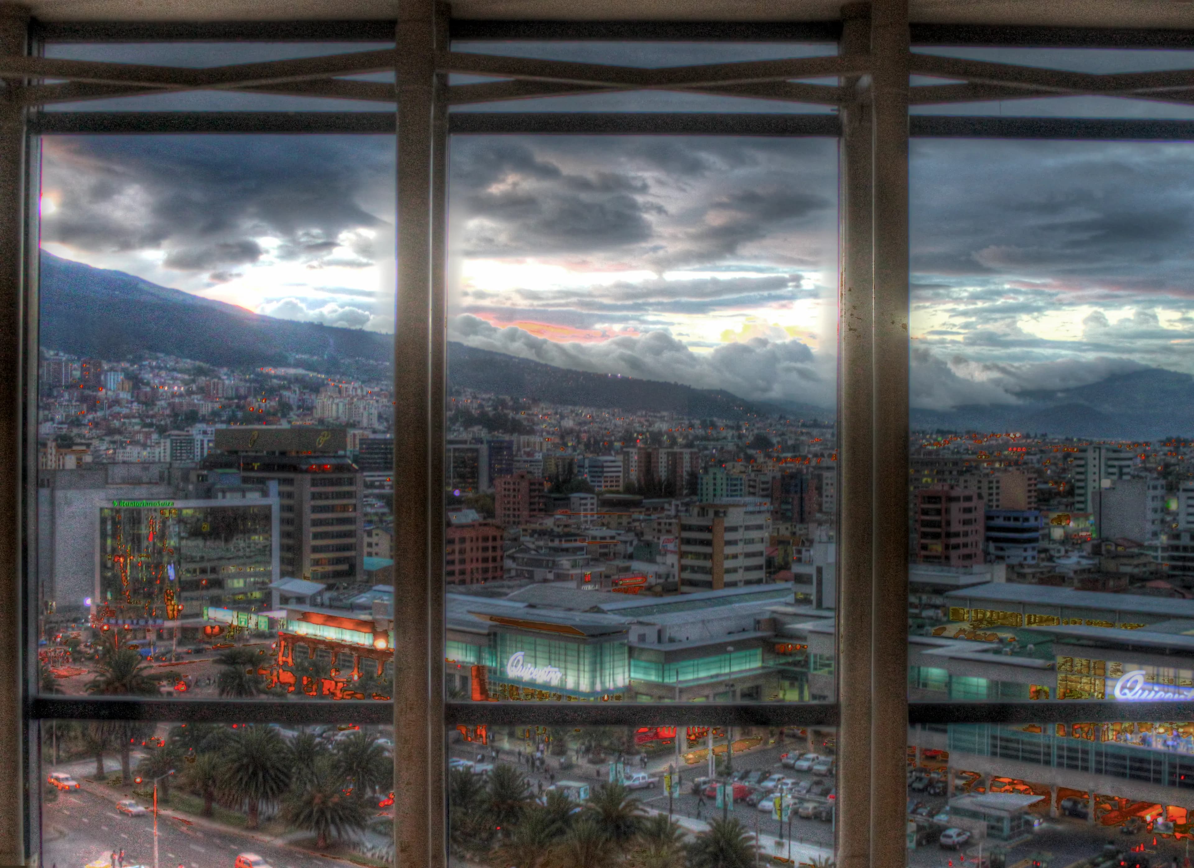 Evening view of Quito, Ecuador, through a barred hotel window with city lights, mountains, and storm clouds, titled The Last Night by Francisco Colón.