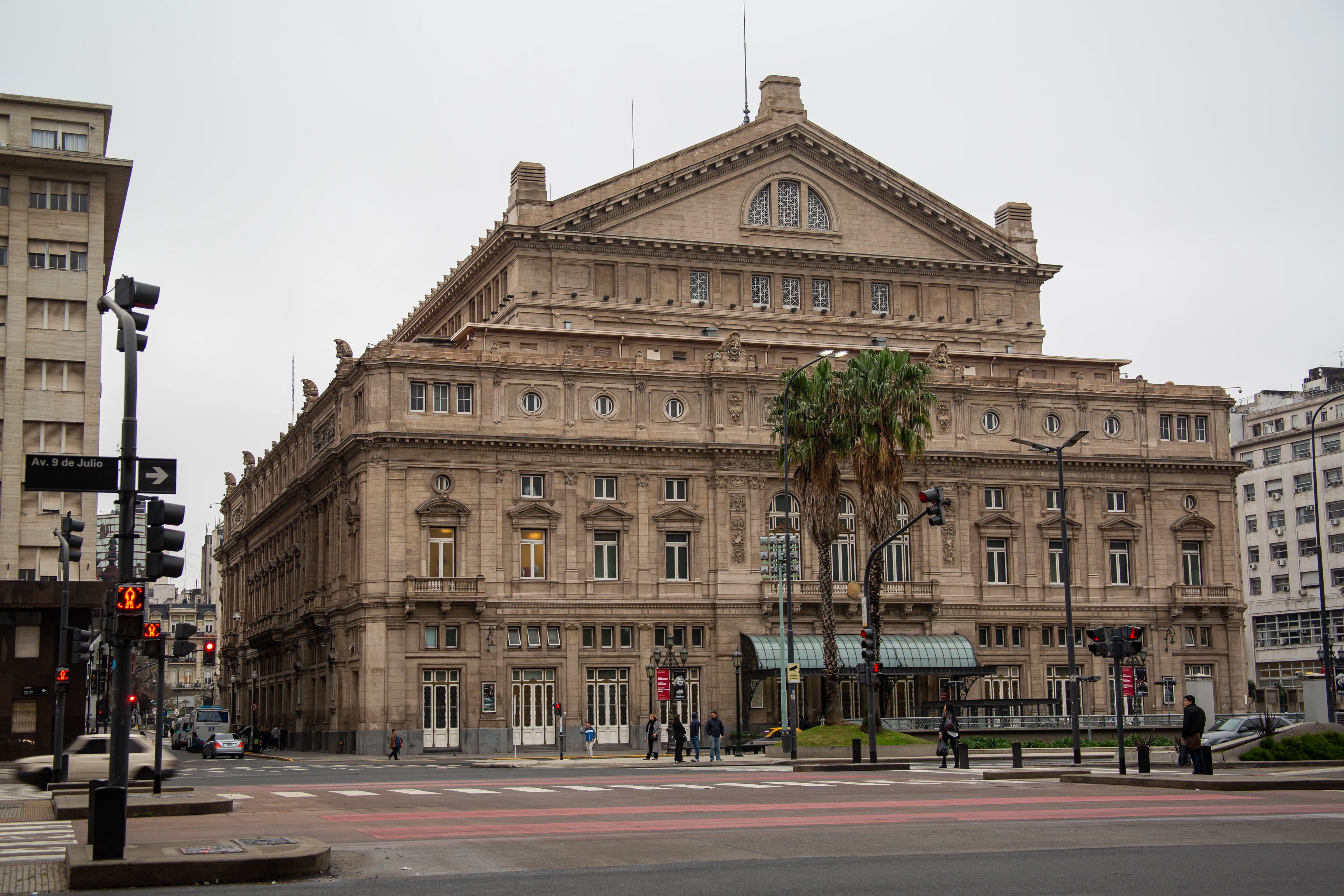 Teatro Colón in Buenos Aires, Argentina, photographed from the surrounding city street with pedestrians, traffic signals, palm trees, and its symmetrical architectural façade visible, titled The Other Colón by Francisco Colón.