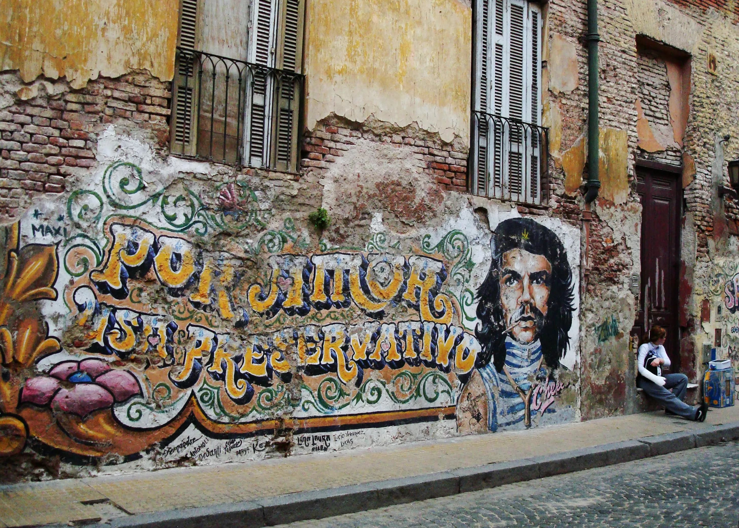 Weathered Buenos Aires wall with an image of Che Guevara, the phrase Por Amor Usa Preservativo, and a young woman seated beside the doorway, titled The Palimpsest by Francisco Colón.