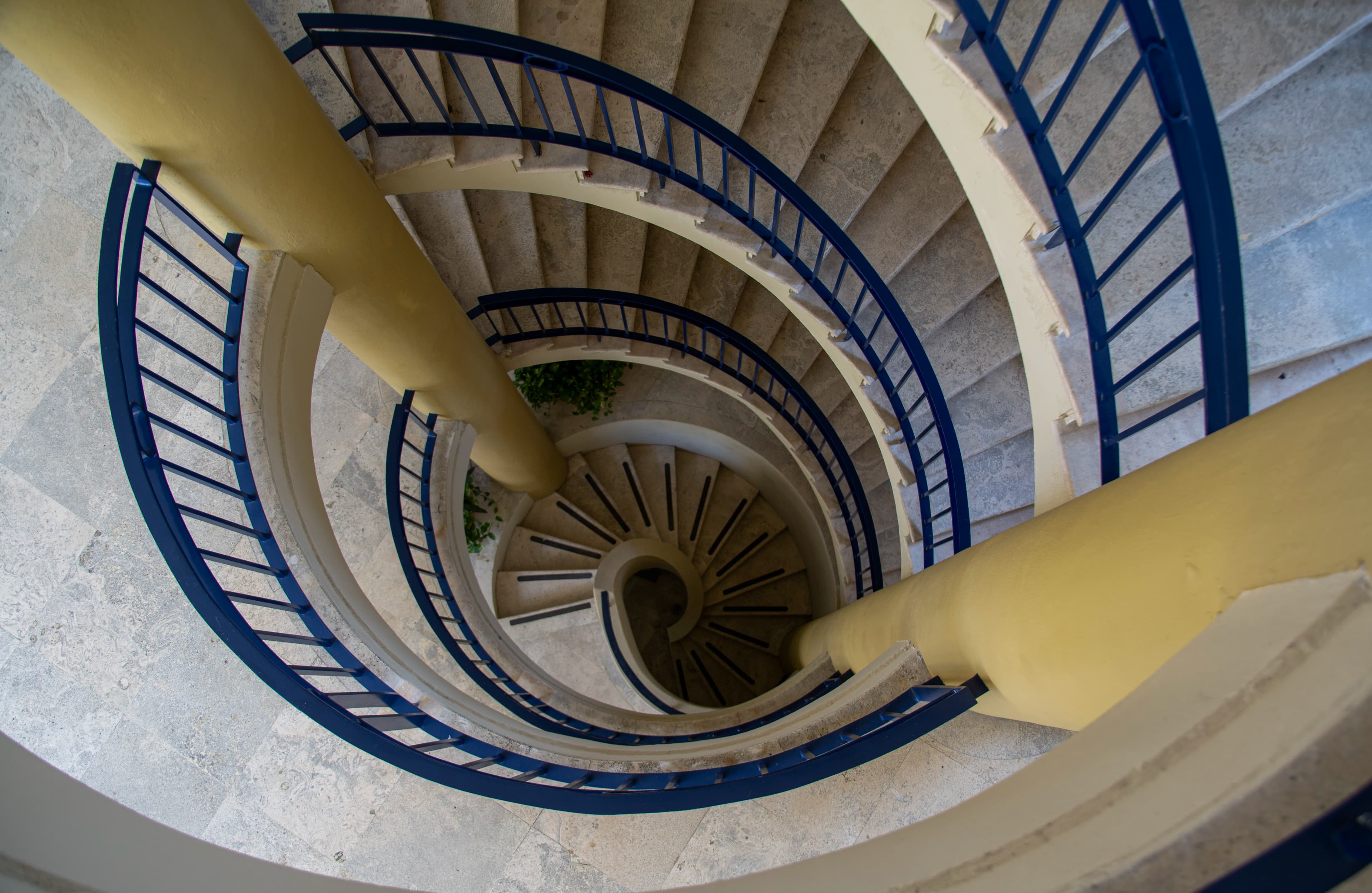 A spiral staircase viewed from above inside a hotel in Cartagena, with blue railings, pale stone steps, yellow columns, and a shadowed circular center.