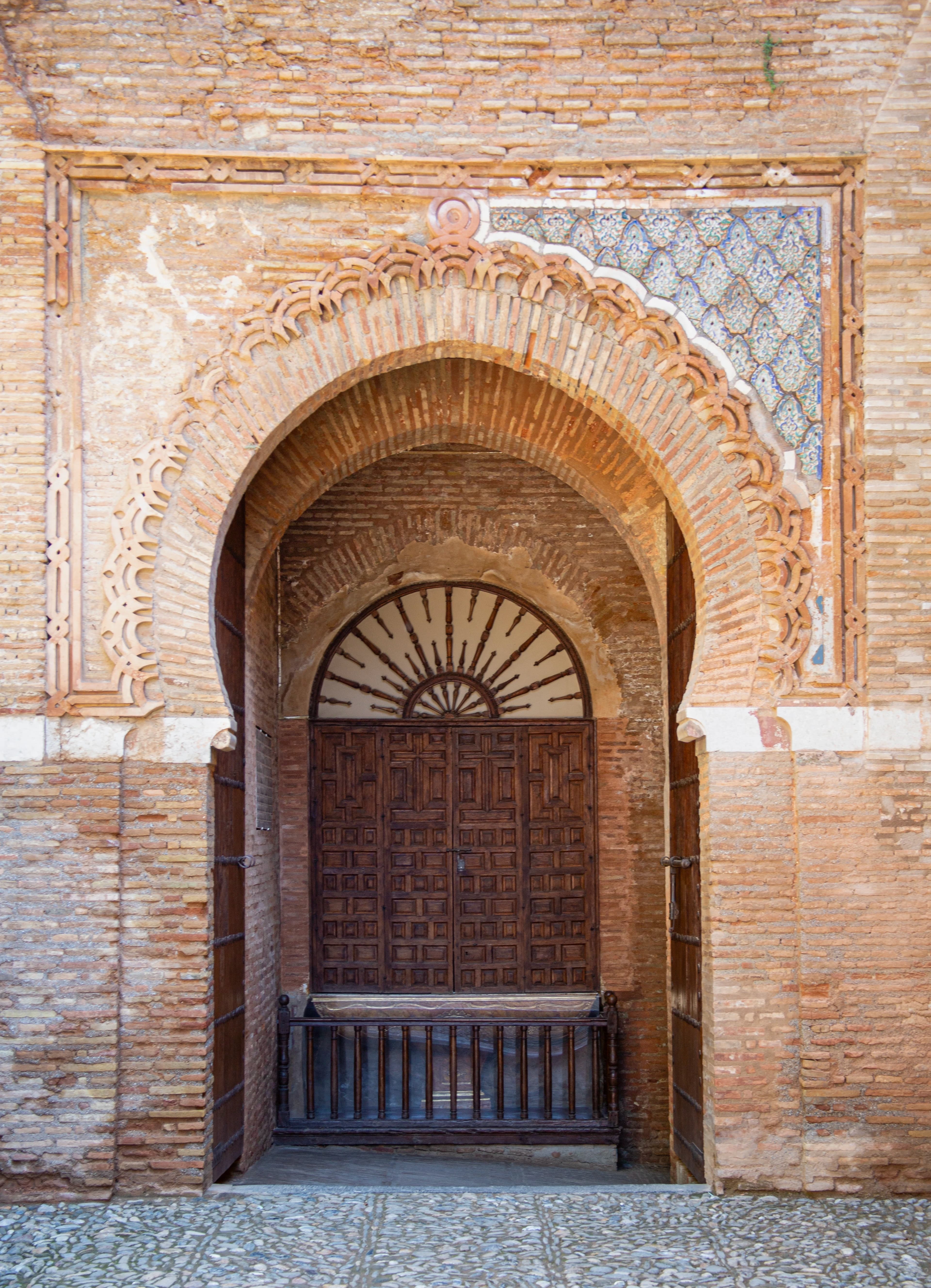 Puerta de la Justicia at the Alhambra in Granada, Spain, showing a monumental horseshoe arch, weathered Nasrid brickwork, surviving blue decorative ornament, recessed wooden doors, and a cobblestone approach.