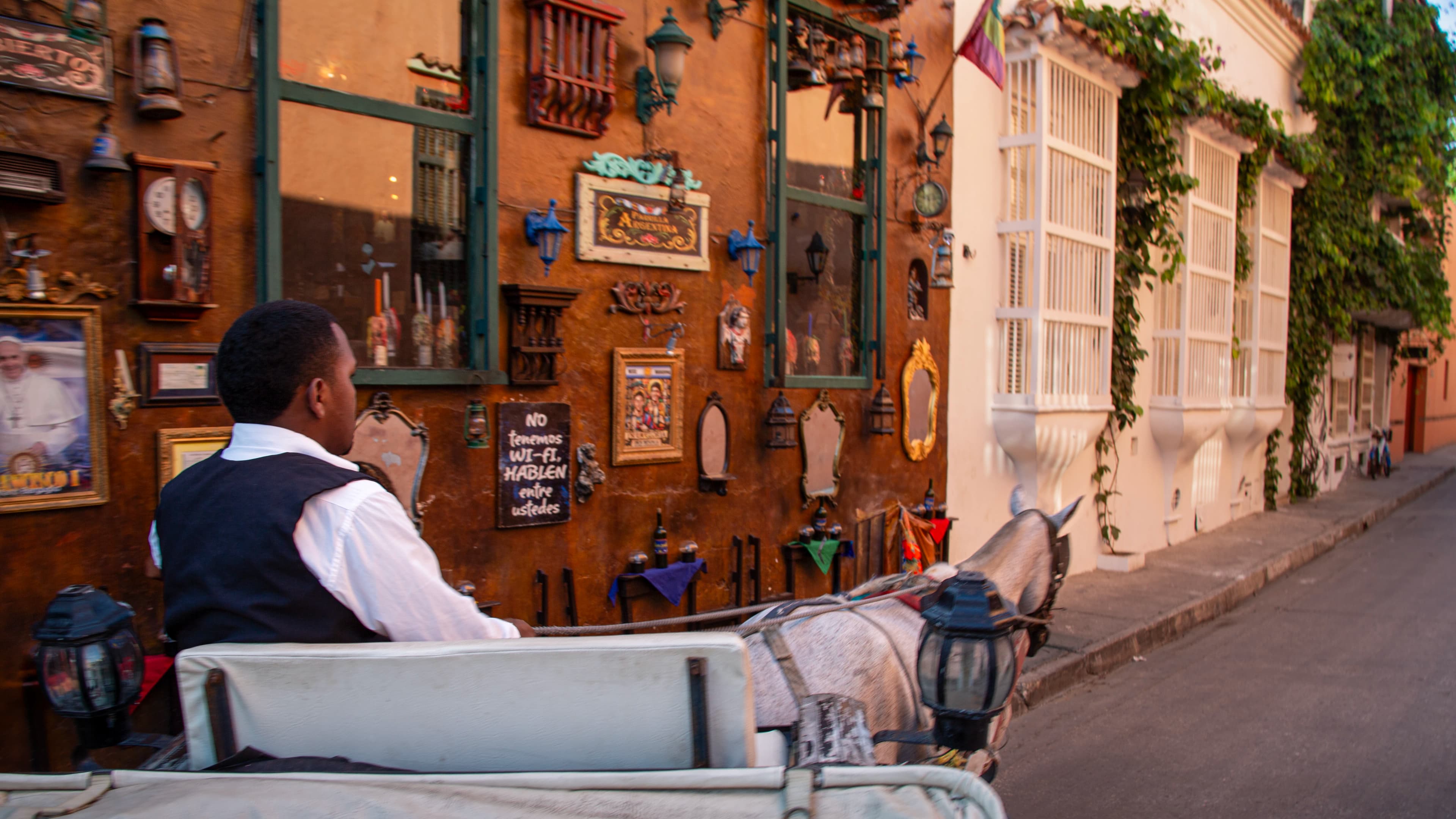 A carriage driver and horse travel past a wall covered with mirrors, clocks, lanterns, framed images, furniture, and a handwritten no Wi-Fi sign in Cartagena's old city.