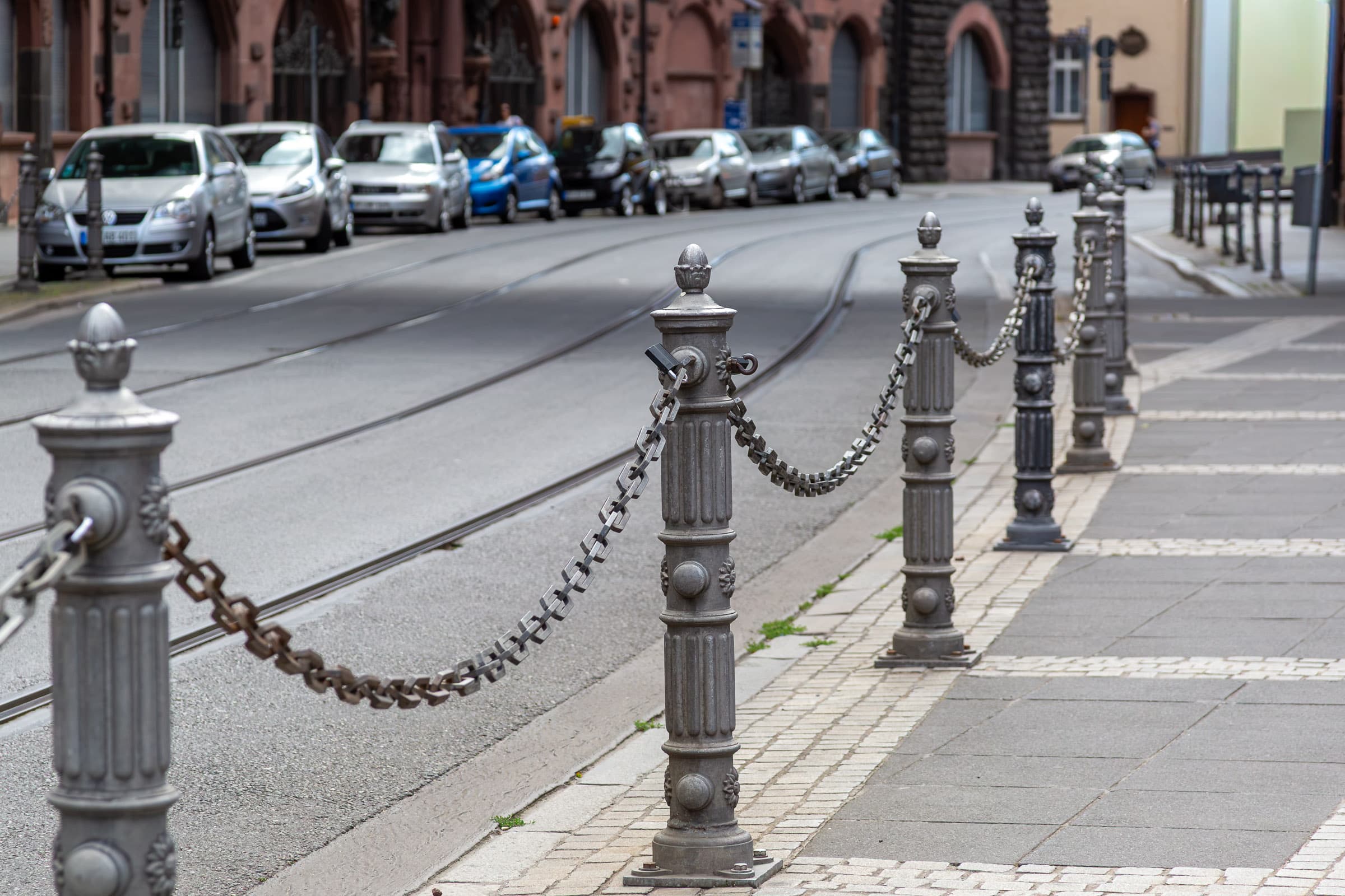 A repeating line of metal posts connected by chains recedes along a Frankfurt street beside tram tracks, parked cars, and urban architecture in What Continues by Francisco Colón.