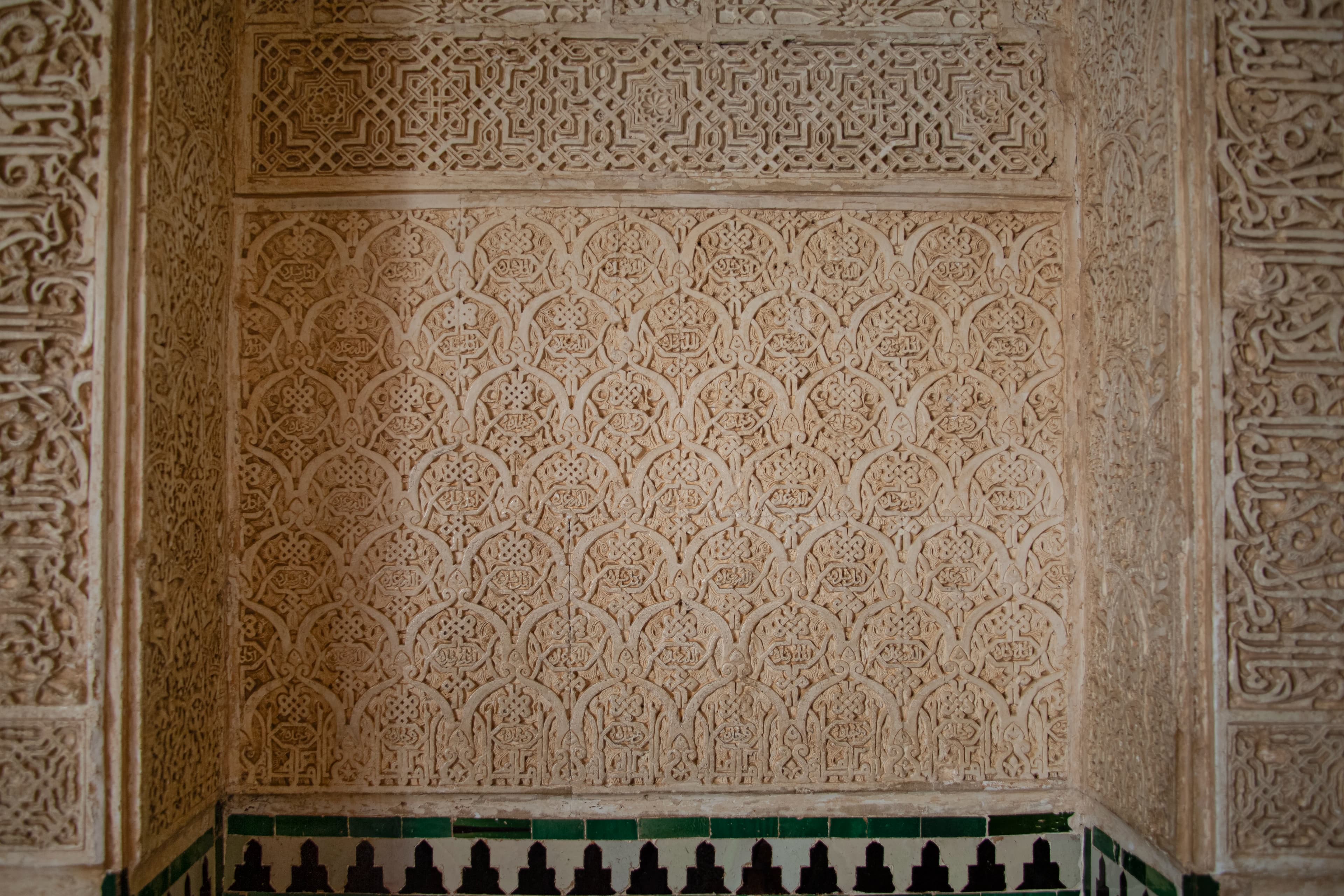Detailed Nasrid plasterwork inside the Alhambra in Granada, Spain, showing repeating geometric and vegetal ornament, Arabic calligraphy, carved architectural borders, and historic tilework.