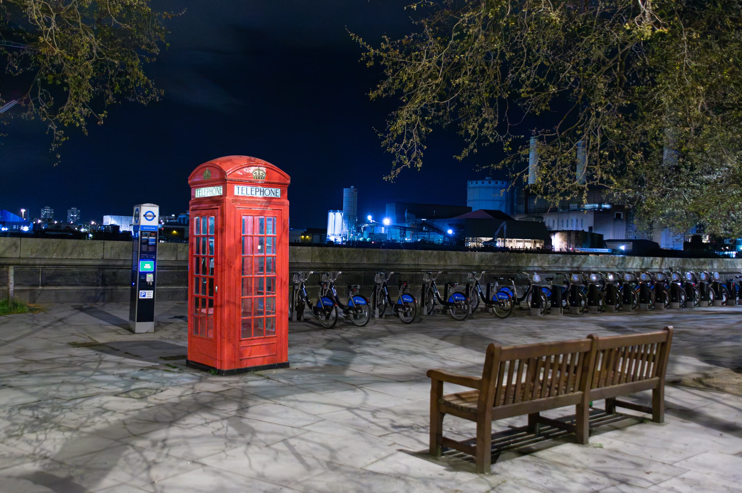 A red telephone box stands near an empty wooden bench and a row of rental bicycles beside the River Thames at night.