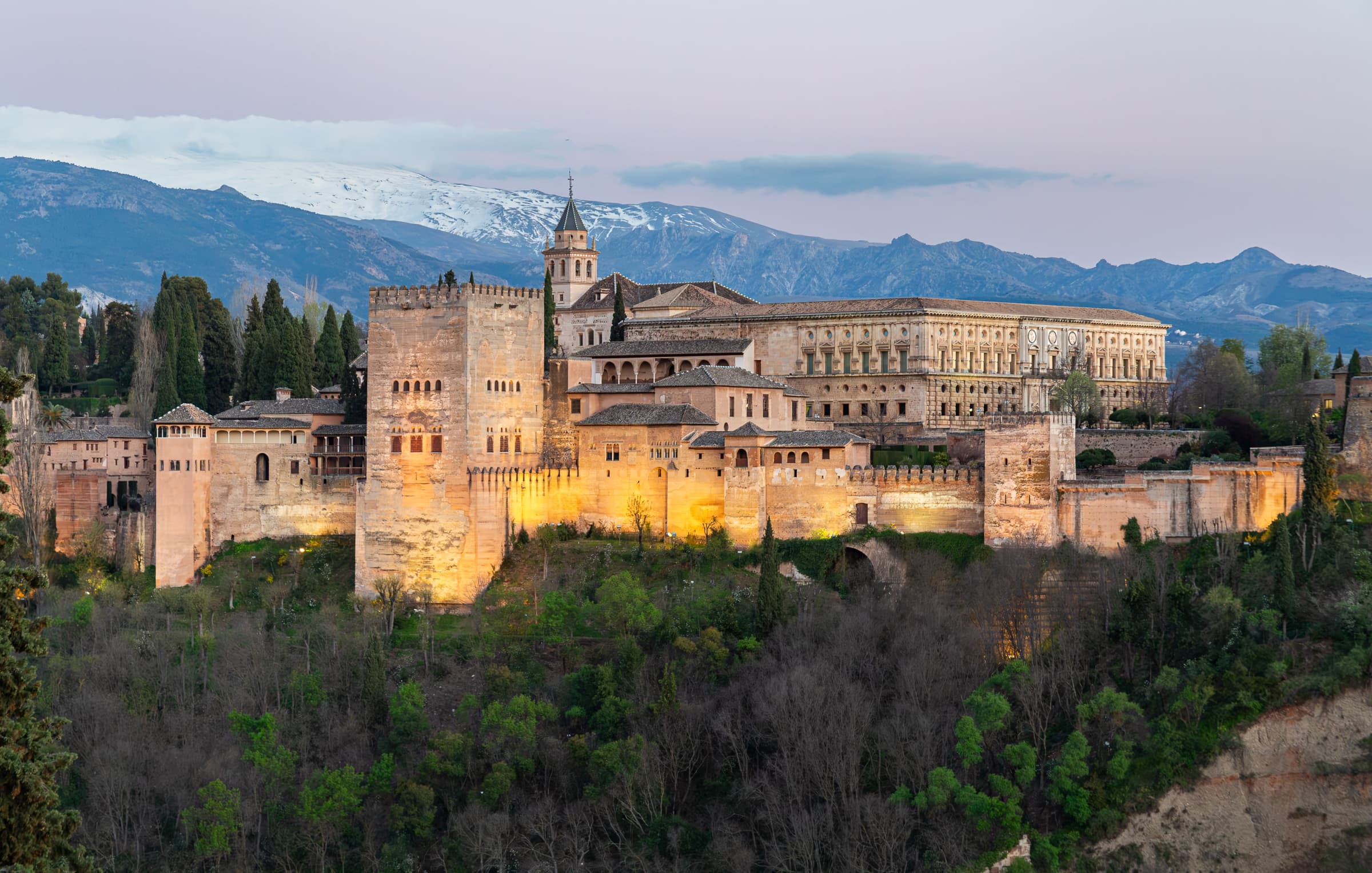 The illuminated Alhambra in Granada, Spain, rises above a wooded hillside with the snow-covered Sierra Nevada behind it in Where Stone Meets the Sierra by Francisco Colón.