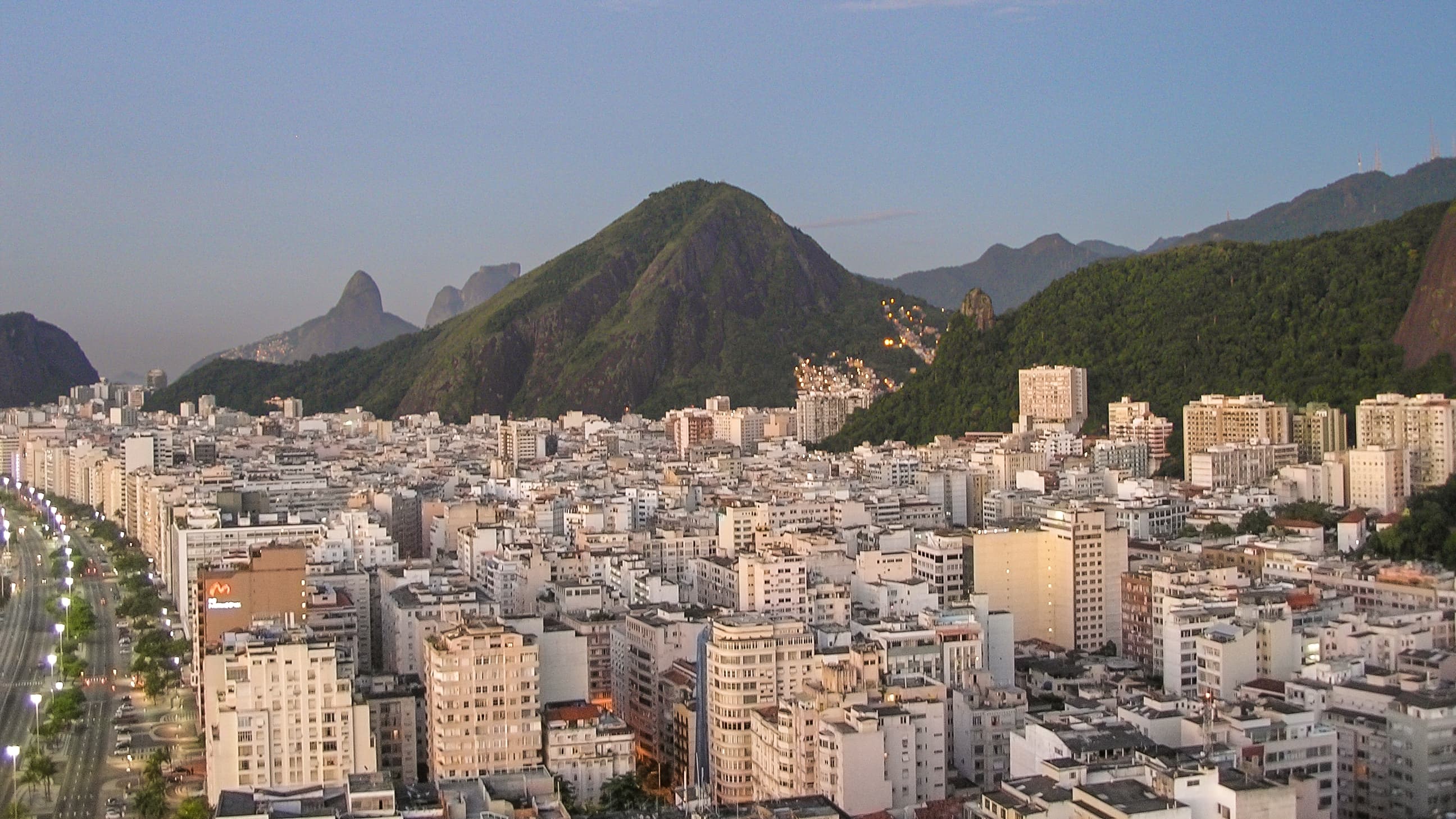 Elevated early-morning view of Copacabana in Rio de Janeiro from a hotel rooftop, showing a brightly illuminated avenue, dense urban buildings, green mountains, rocky peaks, and scattered hillside lights.