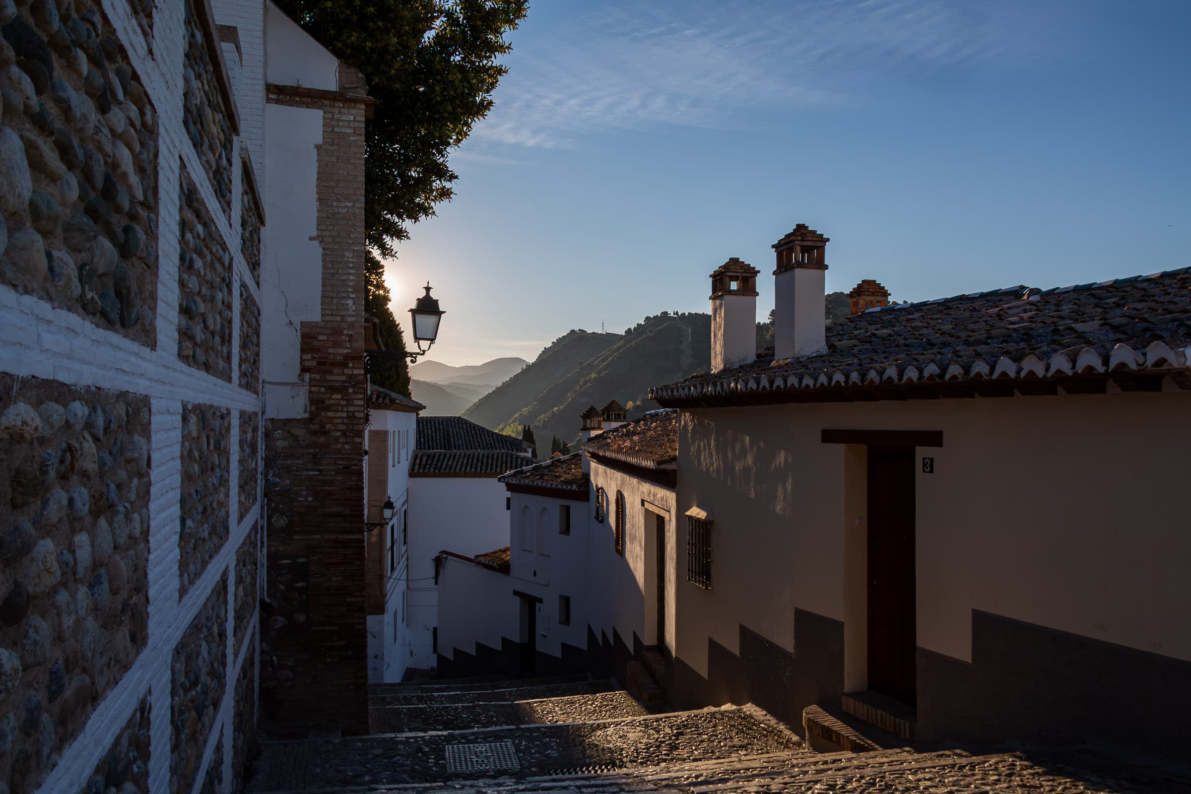 A narrow historic street in Granada, Spain, descending between whitewashed and stone buildings toward mountains illuminated by morning light.