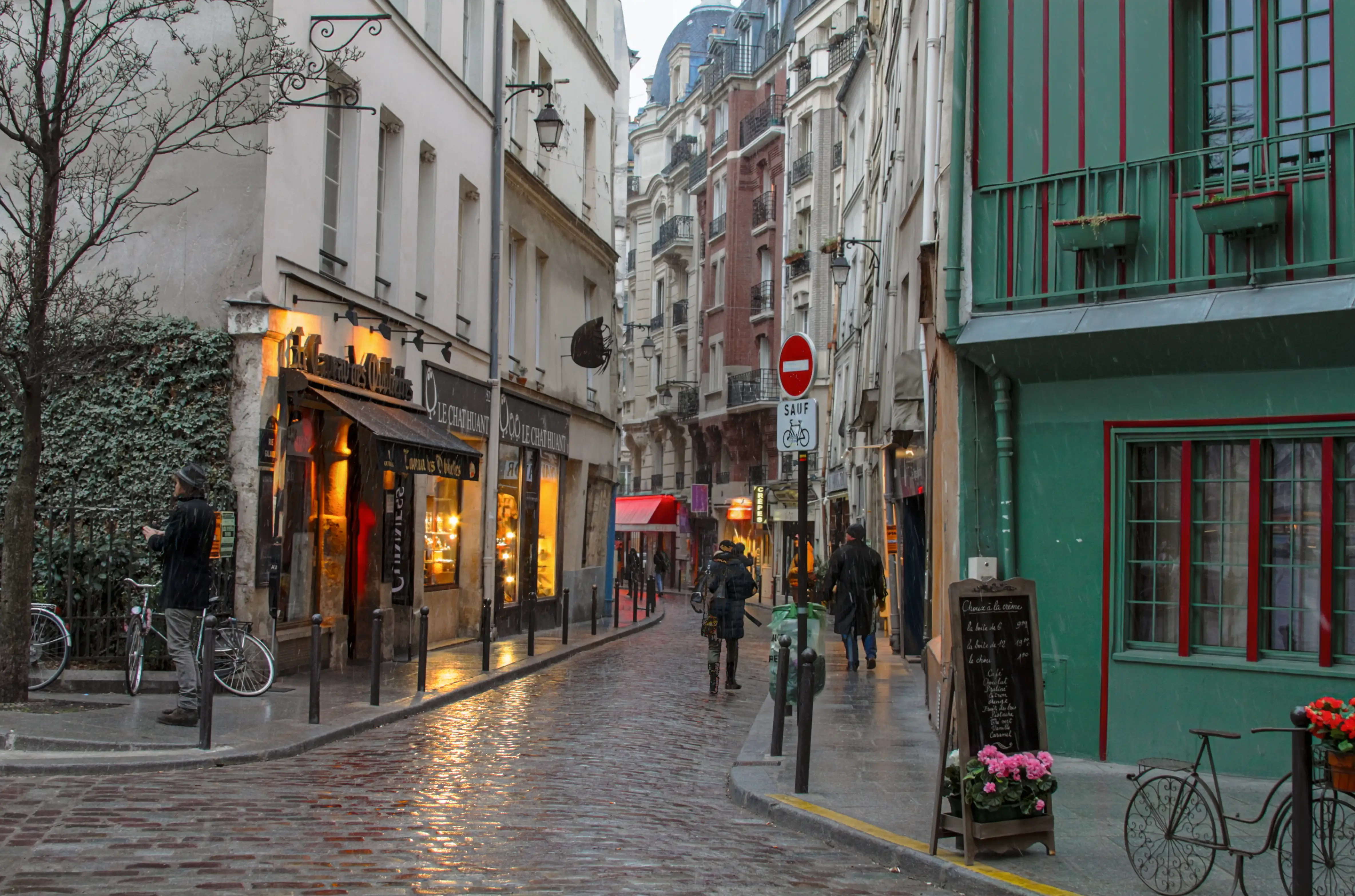 A narrow rain-soaked street in Paris curves out of sight between buildings as pedestrians walk away from the camera and warm restaurant lights reflect across wet cobblestones.