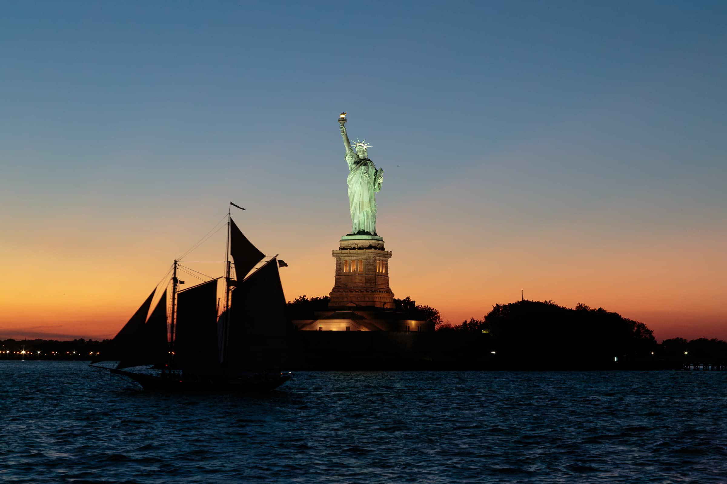 A dark sailboat crosses New York Harbor at dusk with the illuminated Statue of Liberty and Liberty Island behind it.