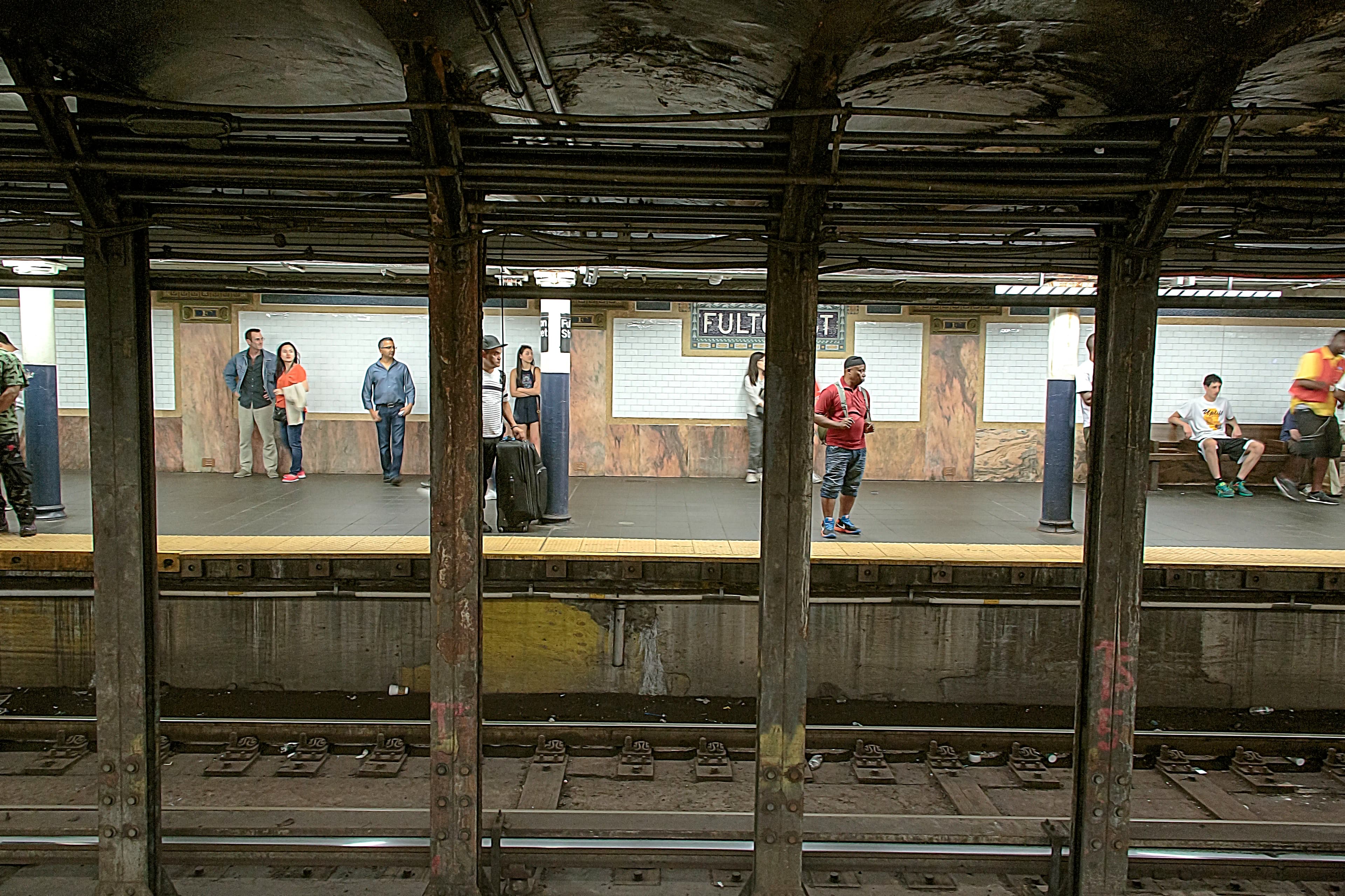 Passengers waiting across the tracks at Fulton Street subway station in New York City, separated visually by steel columns, titled Between Stops by Francisco Colón.