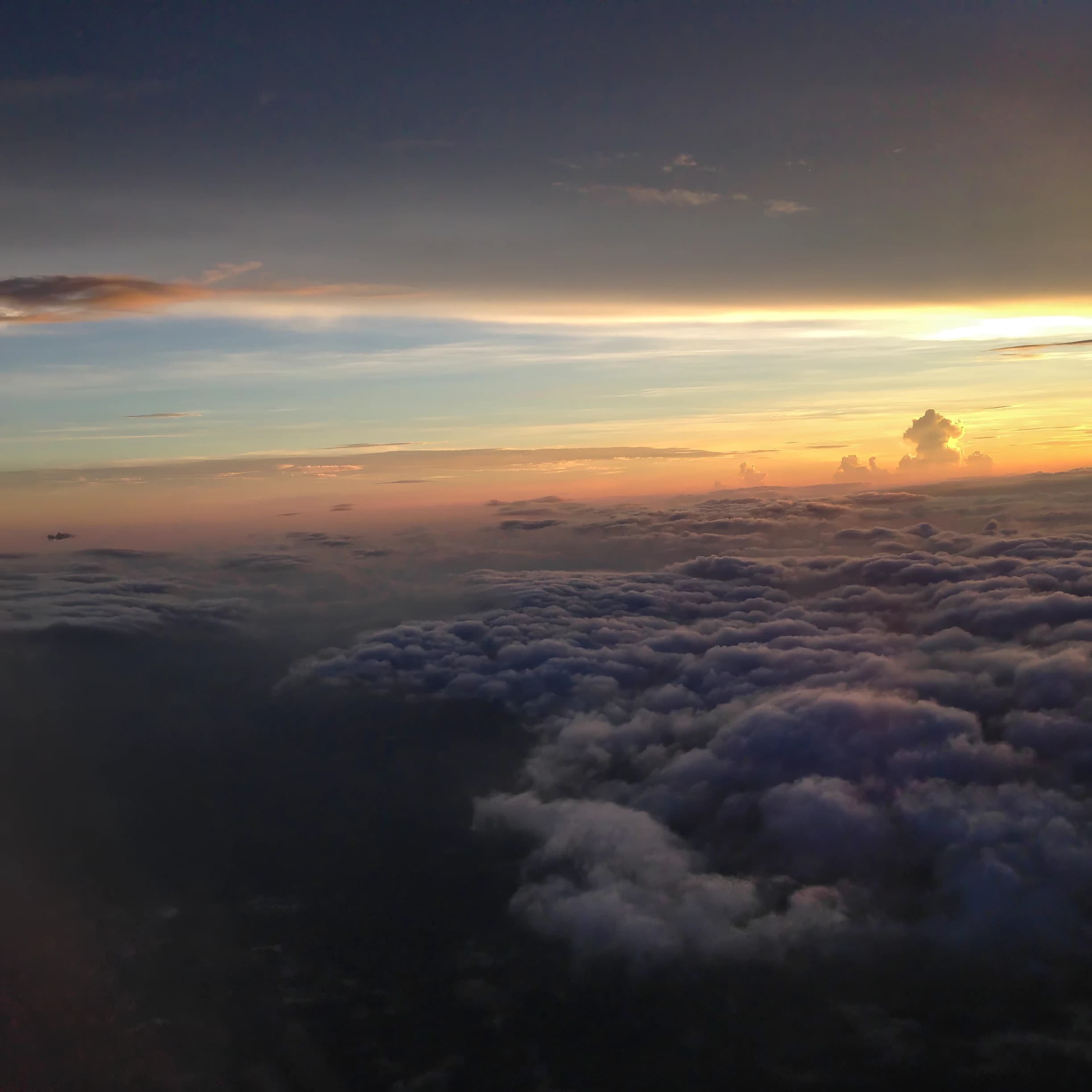 A vast cloudscape photographed from an airplane during a flight to Jamaica, with rolling clouds below, a dark blue-gray sky above, and a narrow luminous horizon of gold, orange, blue, and violet between them.