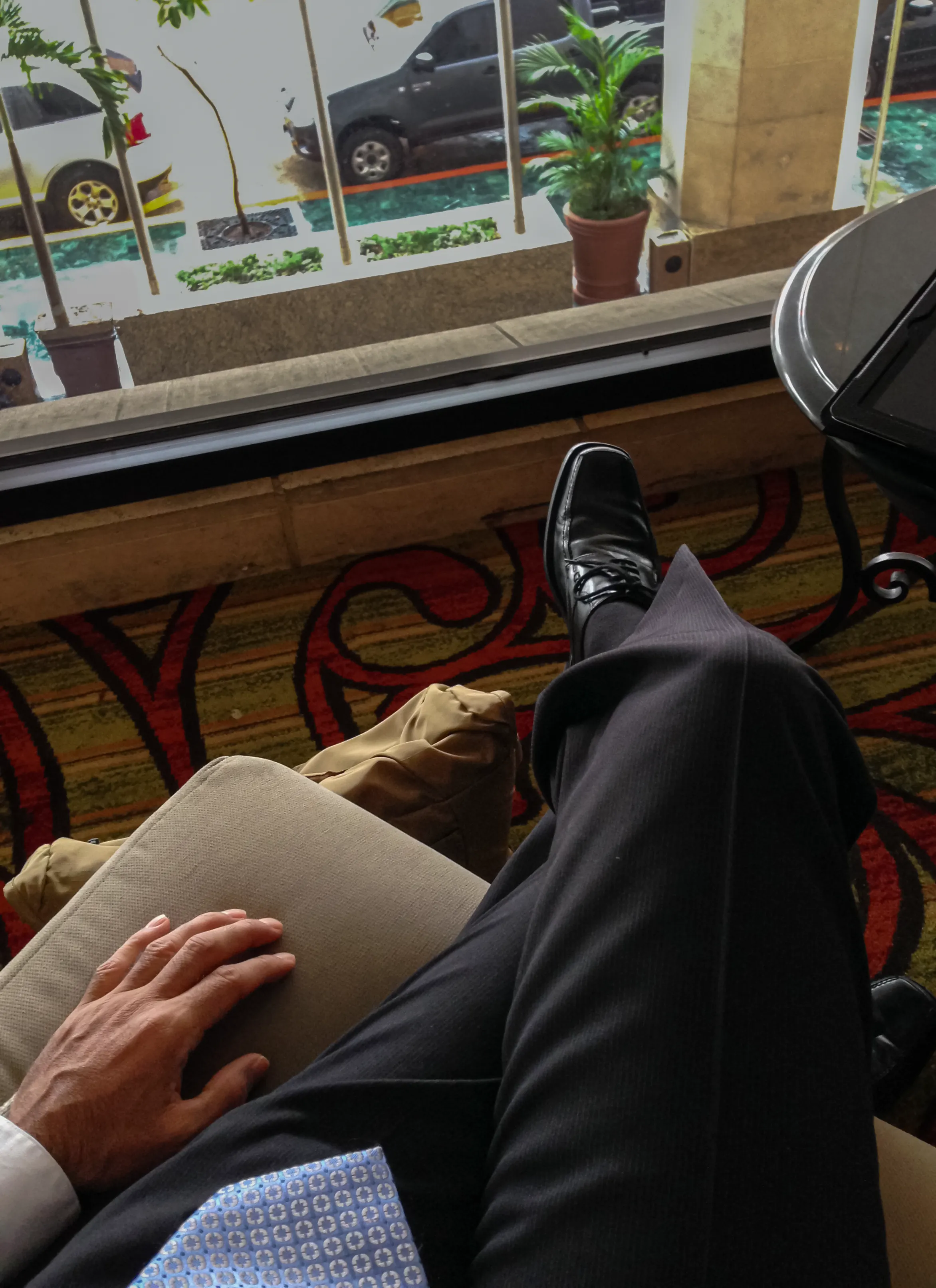 First-person view of Francisco Colón seated in a Marriott hotel lobby in El Salvador wearing business attire and dress shoes with his work bag beside him while waiting for transportation to a customer meeting, titled Ready to Go.