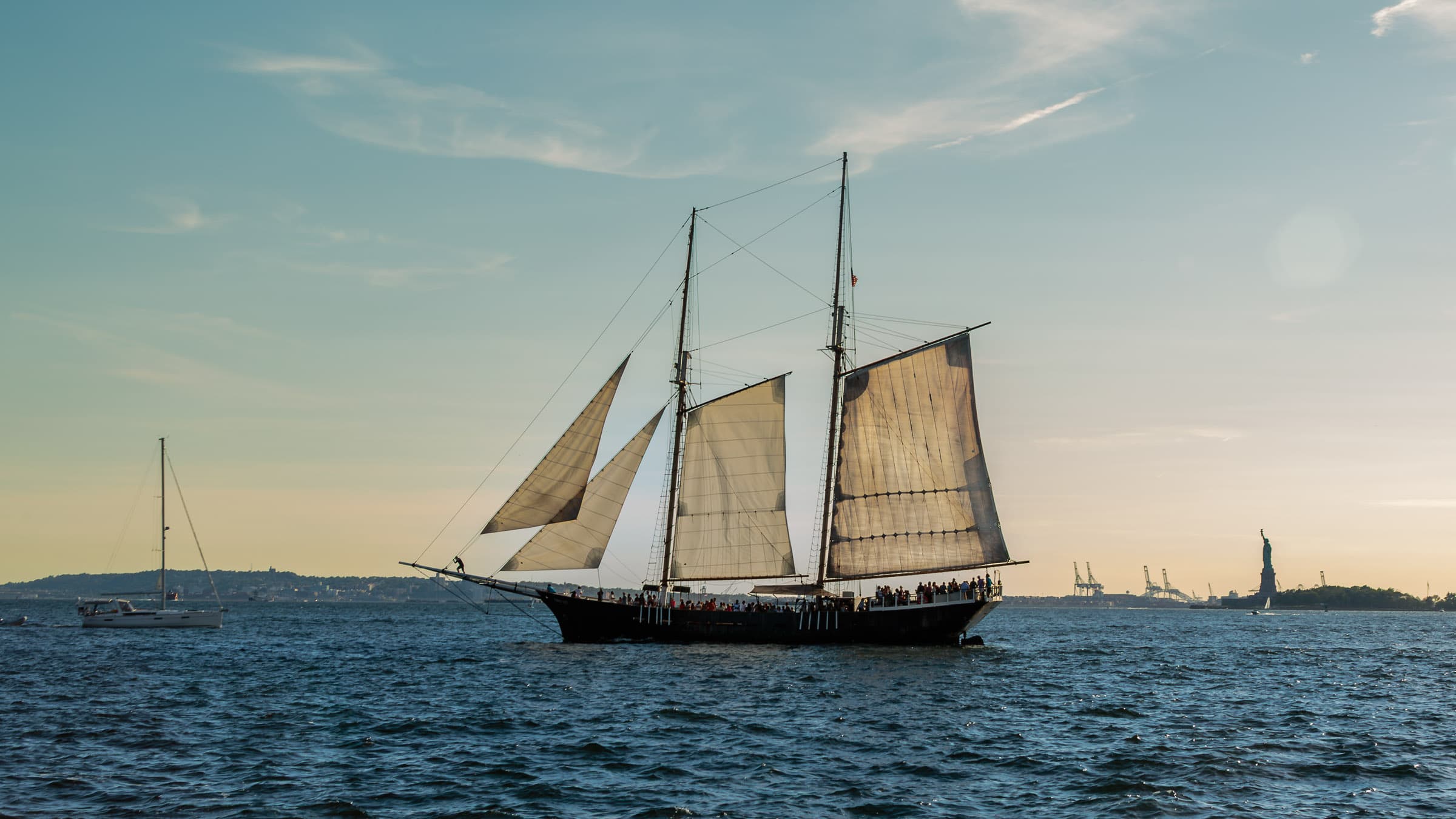 Traditional schooner sailing across New York Harbor with a smaller sailboat to the left and the Statue of Liberty in the distance to the right, photographed by Francisco Colón.