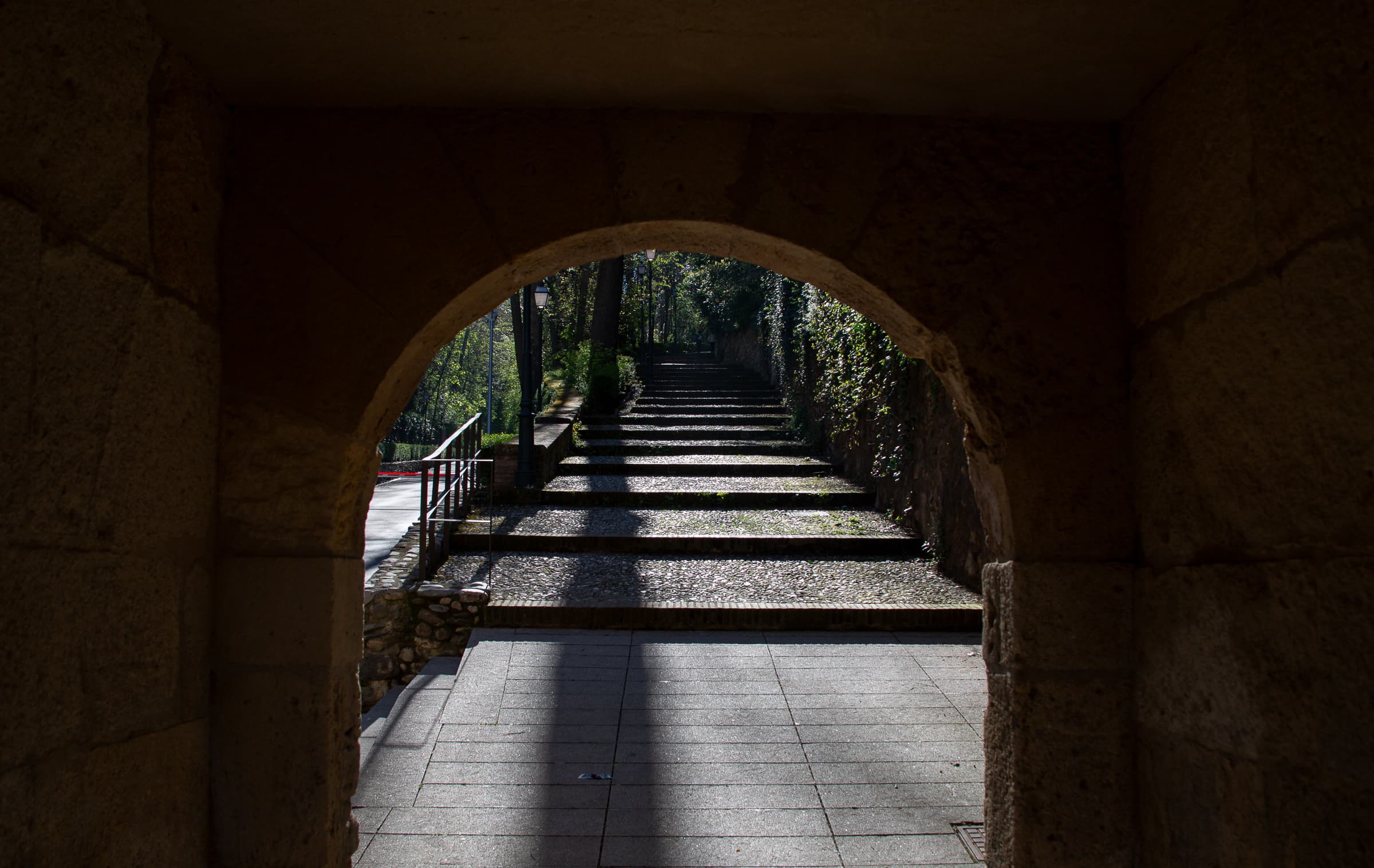 A dark stone archway in Granada, Spain, framing a staircase rising through alternating bands of morning sunlight and shadow toward surrounding greenery.