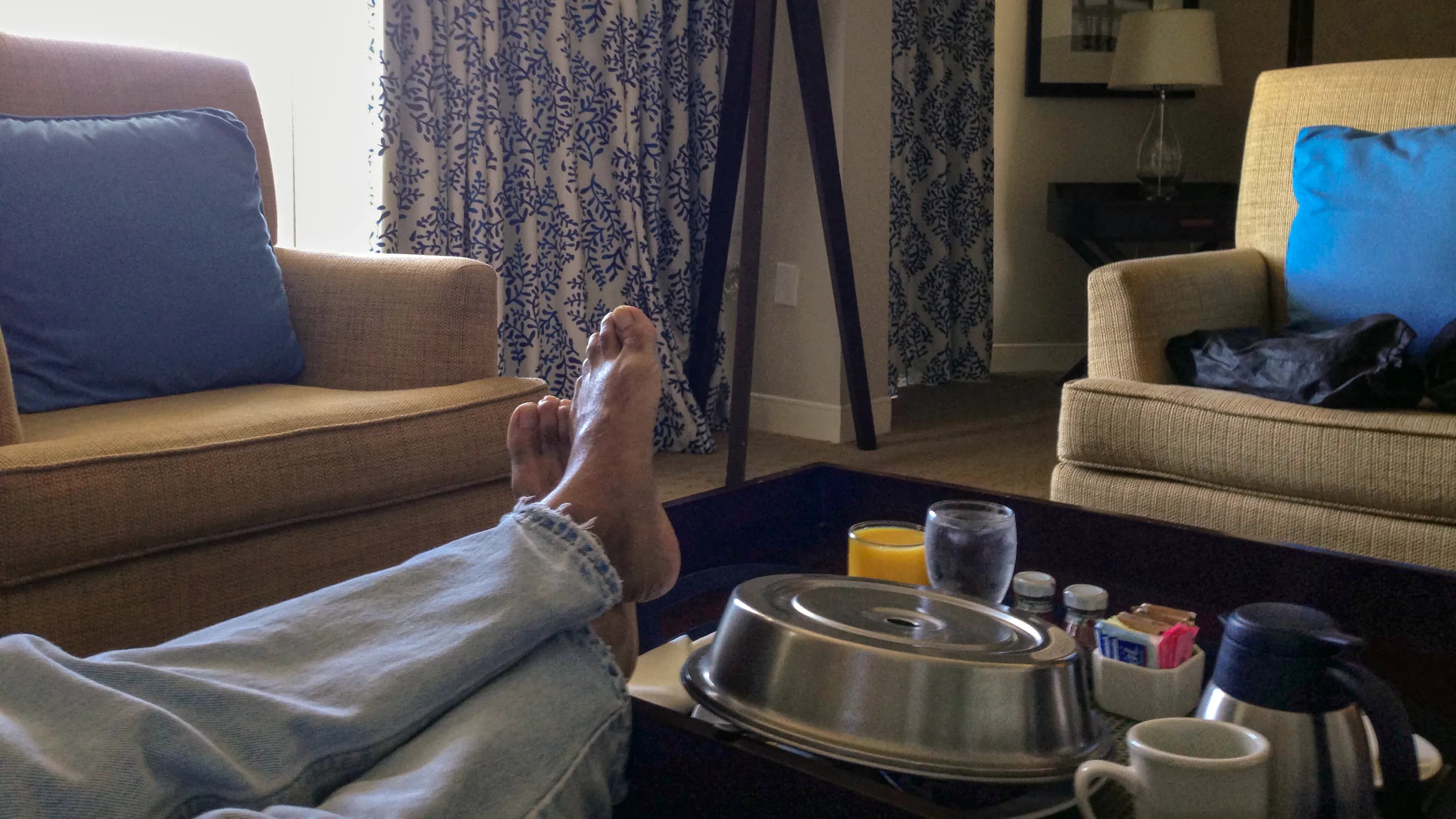 First-person view of a traveler resting with his feet elevated beside a room-service breakfast in a Sheraton hotel room in the Bahamas, titled The Waiting Room by Francisco Colón.