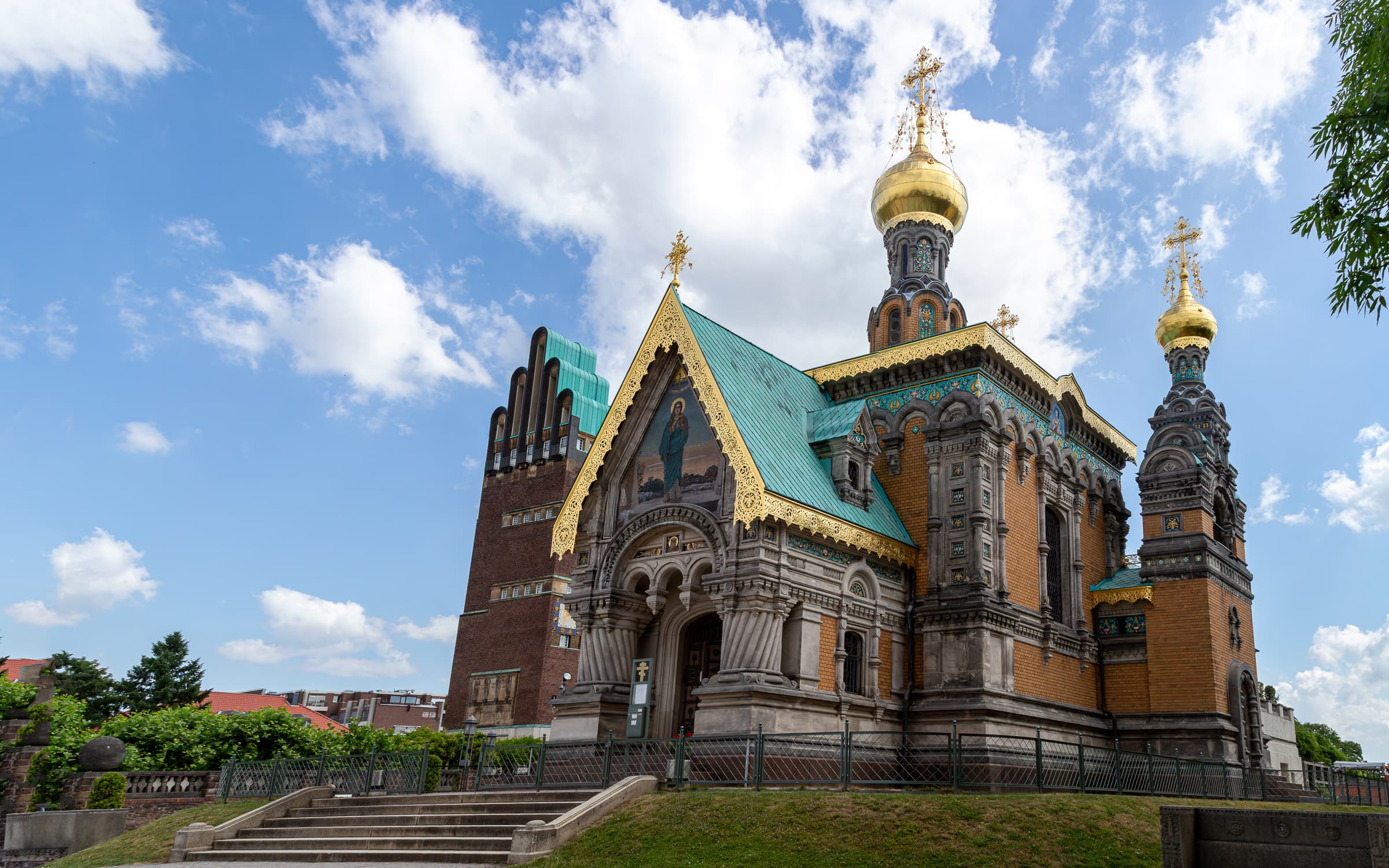 Panoramic view of the Russian Chapel of St. Mary Magdalene at Mathildenhöhe in Darmstadt, Germany, with gilded onion domes, turquoise roofing, ornate mosaics, and the neighboring Wedding Tower beneath a blue and cloud-filled sky.