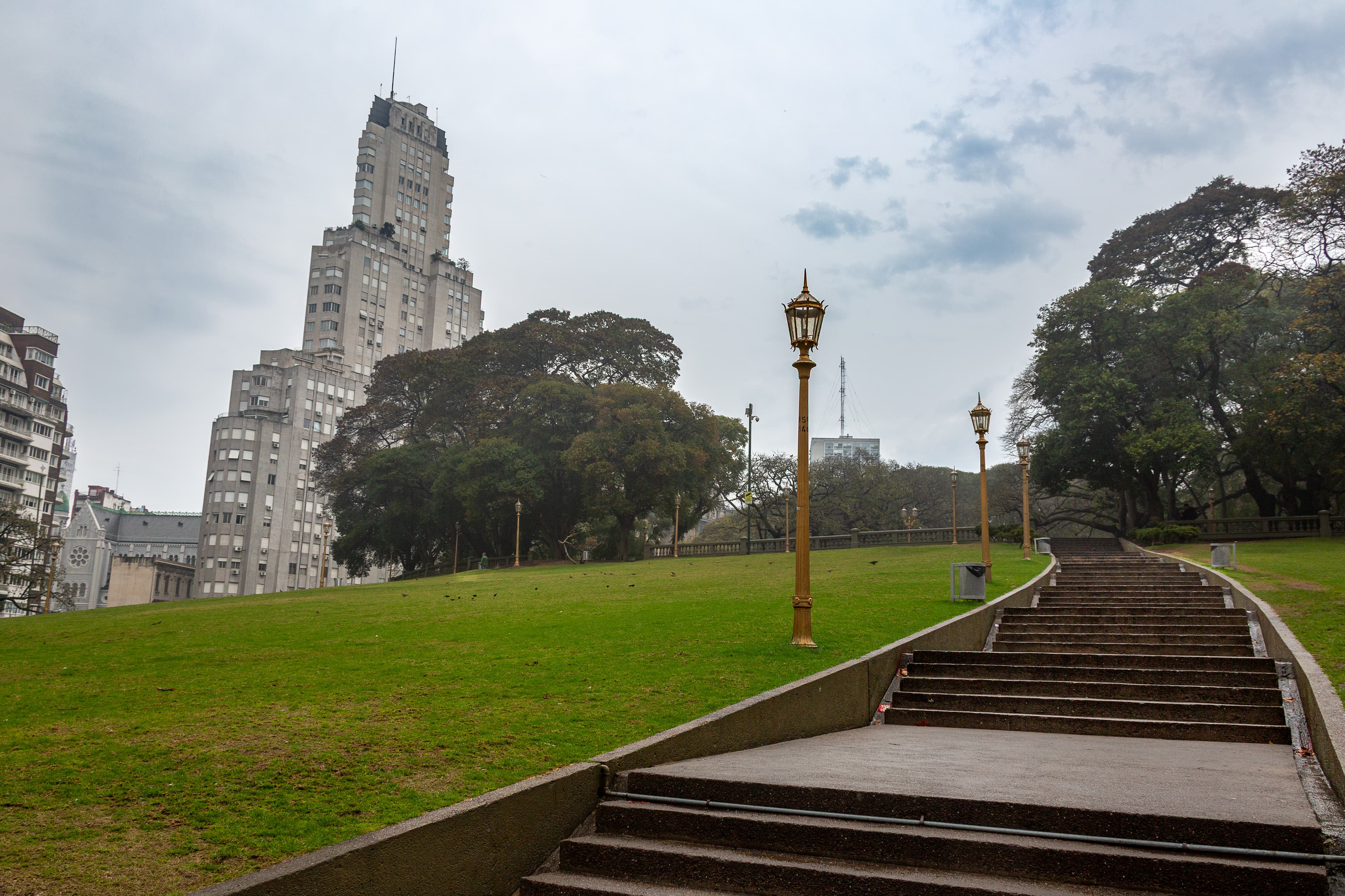 Early morning view of Plaza San Martín in Buenos Aires with a staircase rising through an empty green lawn, repeating lampposts, mature trees, birds and surrounding city architecture under an overcast sky.