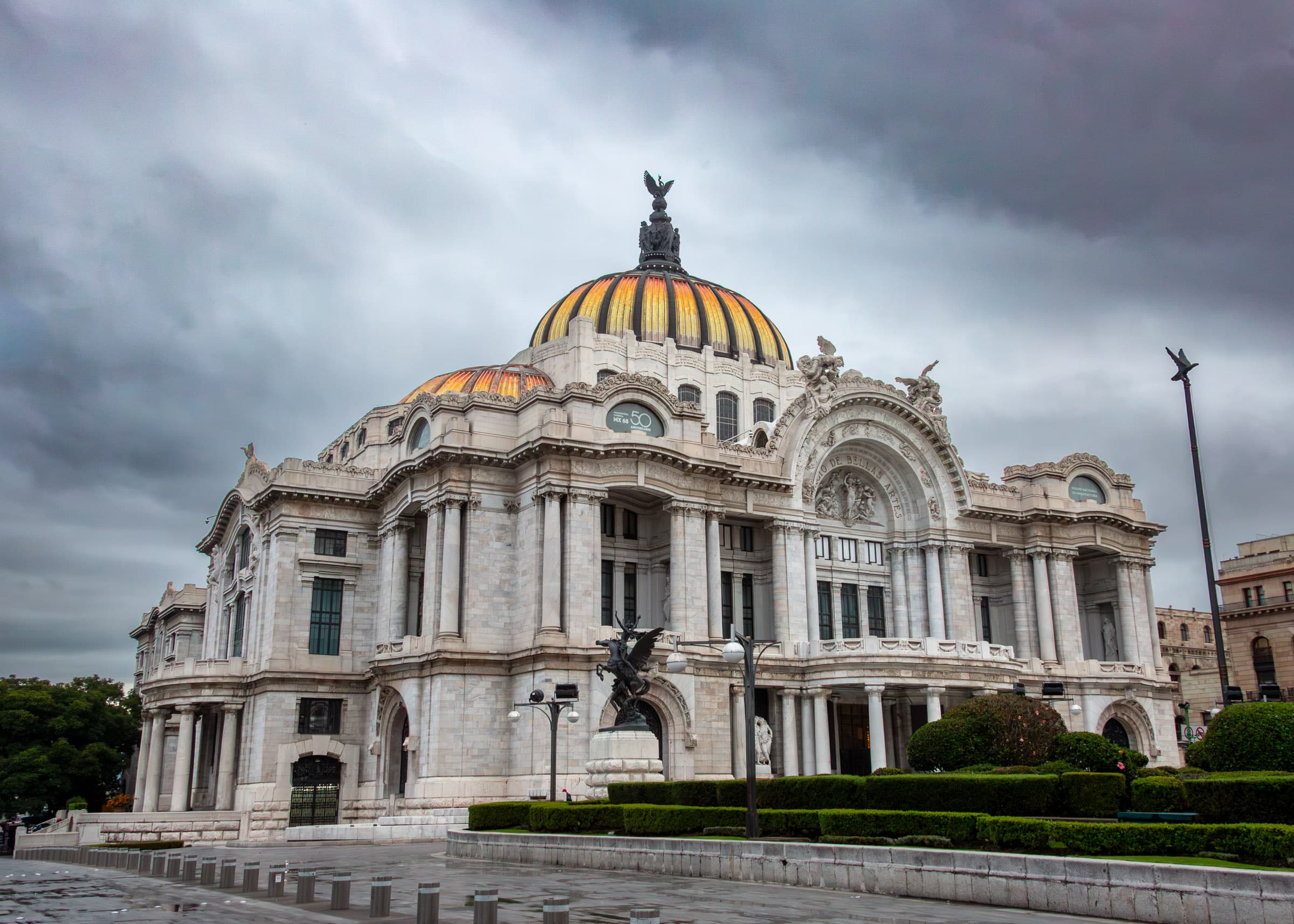 Palacio de Bellas Artes in Mexico City beneath a dramatic cloudy morning sky, with its warm-colored dome rising above the pale stone architecture, photographed by Francisco Colón.