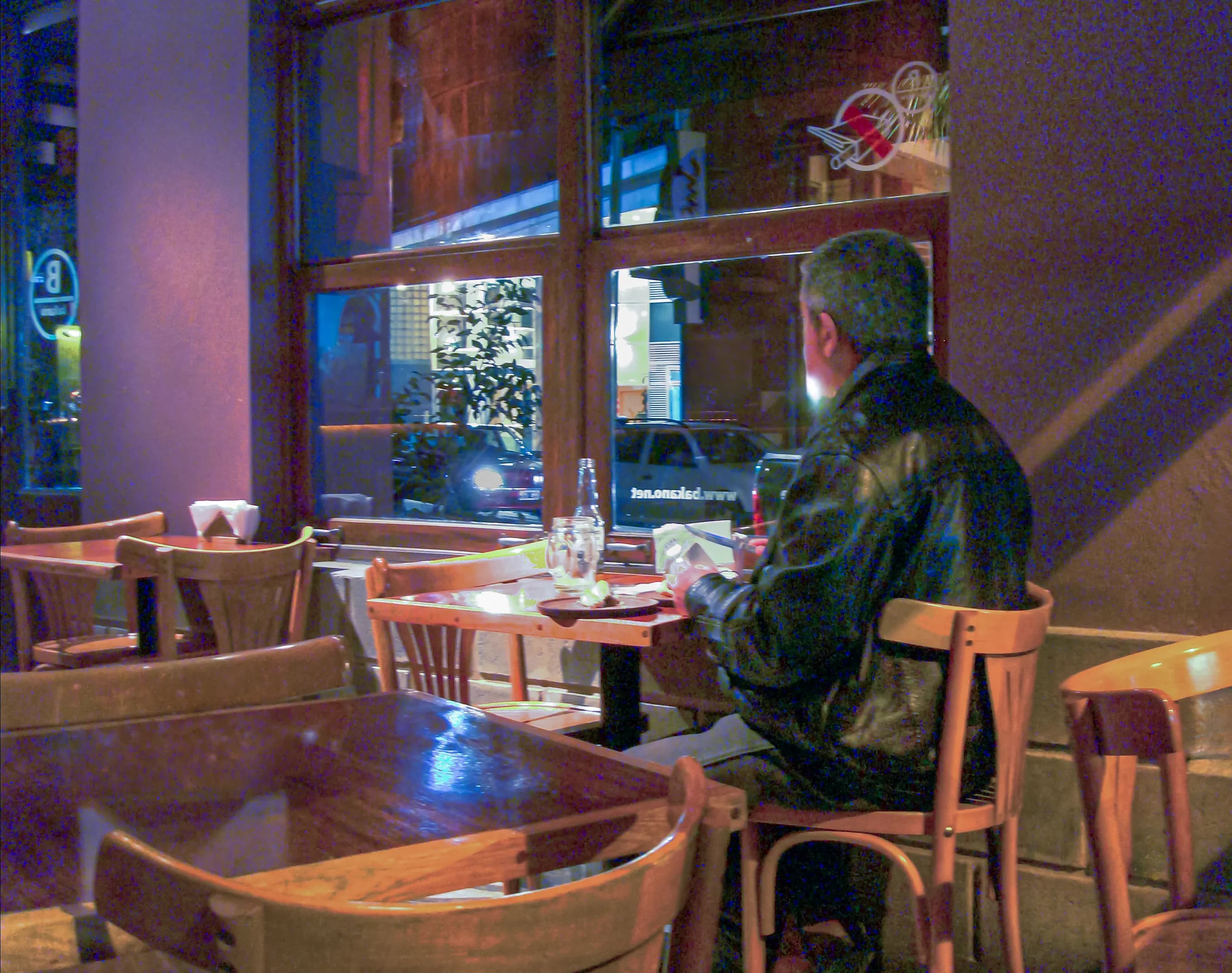 Man seated alone beside a restaurant window in Palermo, Buenos Aires, with an untouched chair across the table, titled Where Thoughts Come to Rest by Francisco Colón.